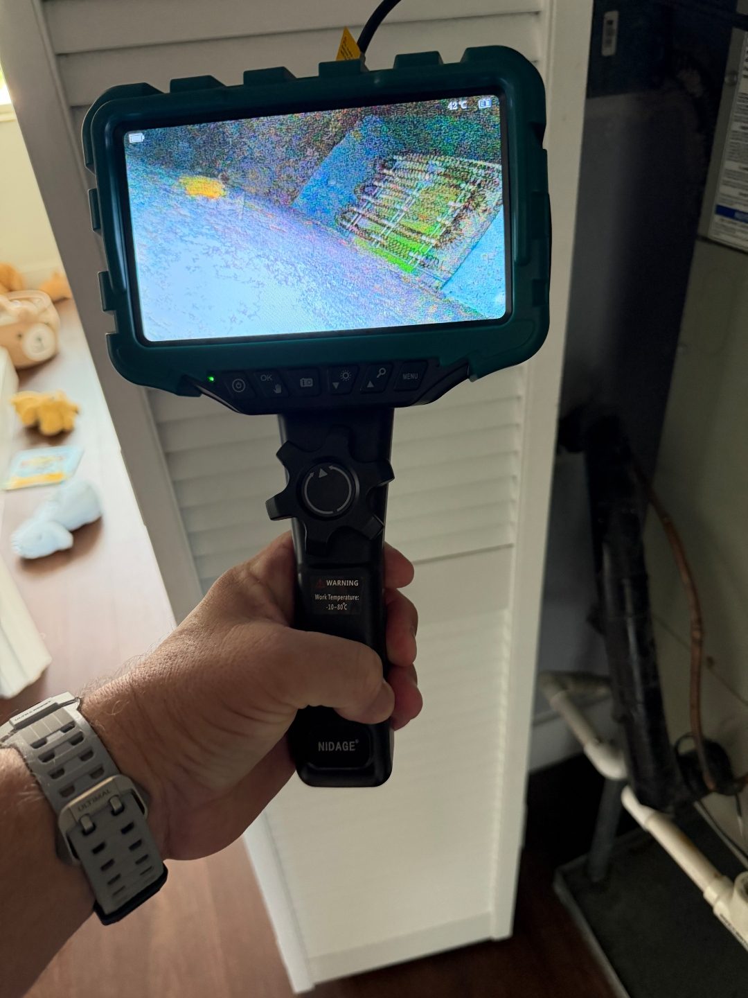 Hand holding a borescope camera in front of a louvered closet door, screen showing a scan with a coil pattern
