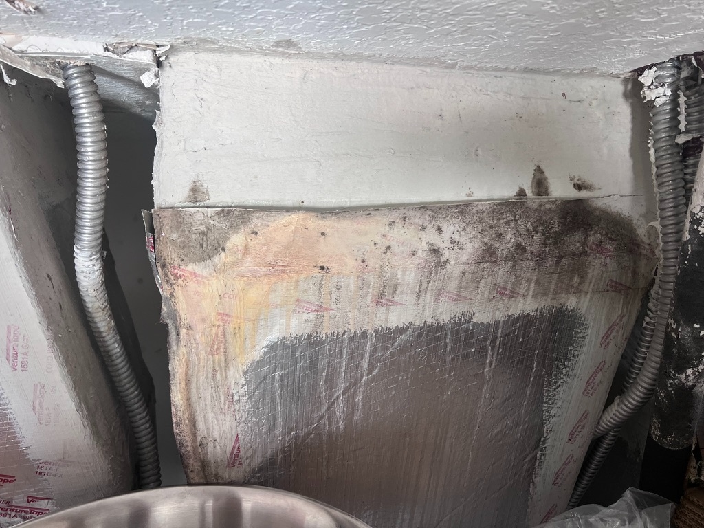 Wide view of an HVAC plenum insulation panel showing dark mold growth along the top seam where the panel meets the ceiling framing