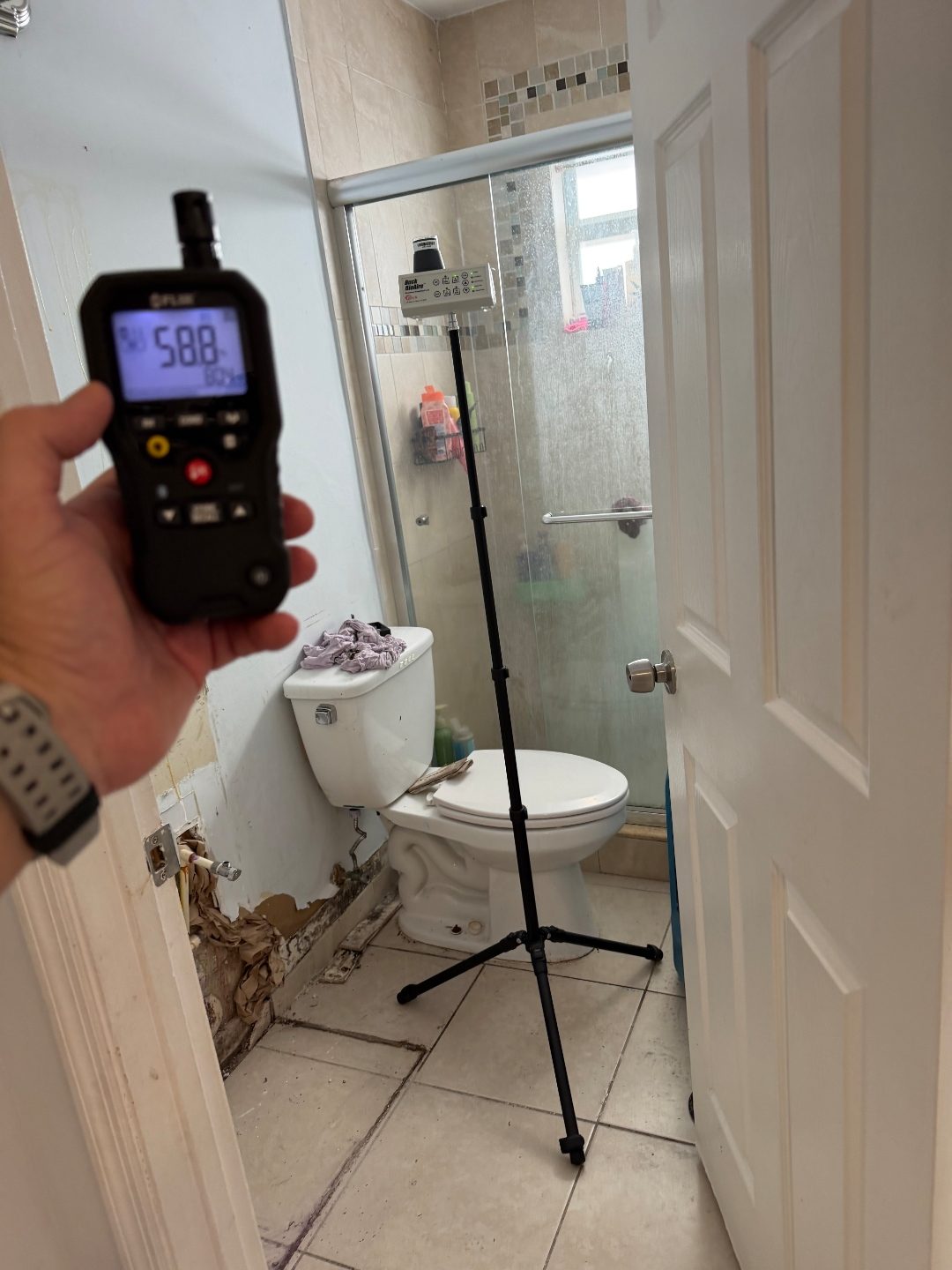 Hand holding a digital moisture meter in a bathroom doorway with a toilet and shower enclosure visible