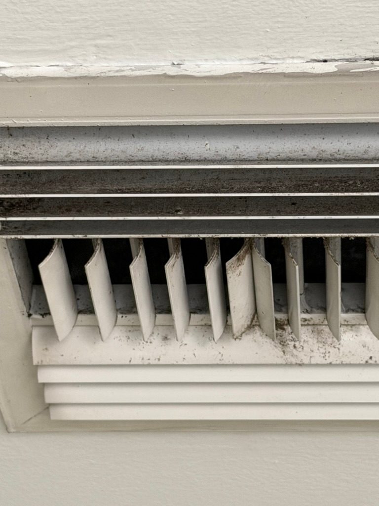 Close-up of a wall mounted return air grille with dust and debris on the louvers