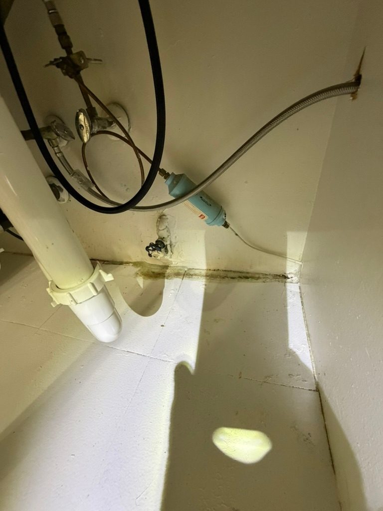 Interior of a sink base cabinet showing supply lines and staining along the wall to floor joint