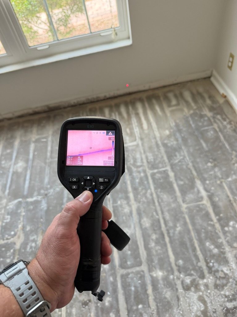 Hand holding a thermal imaging camera displaying a scan near a window wall in a room with the flooring removed